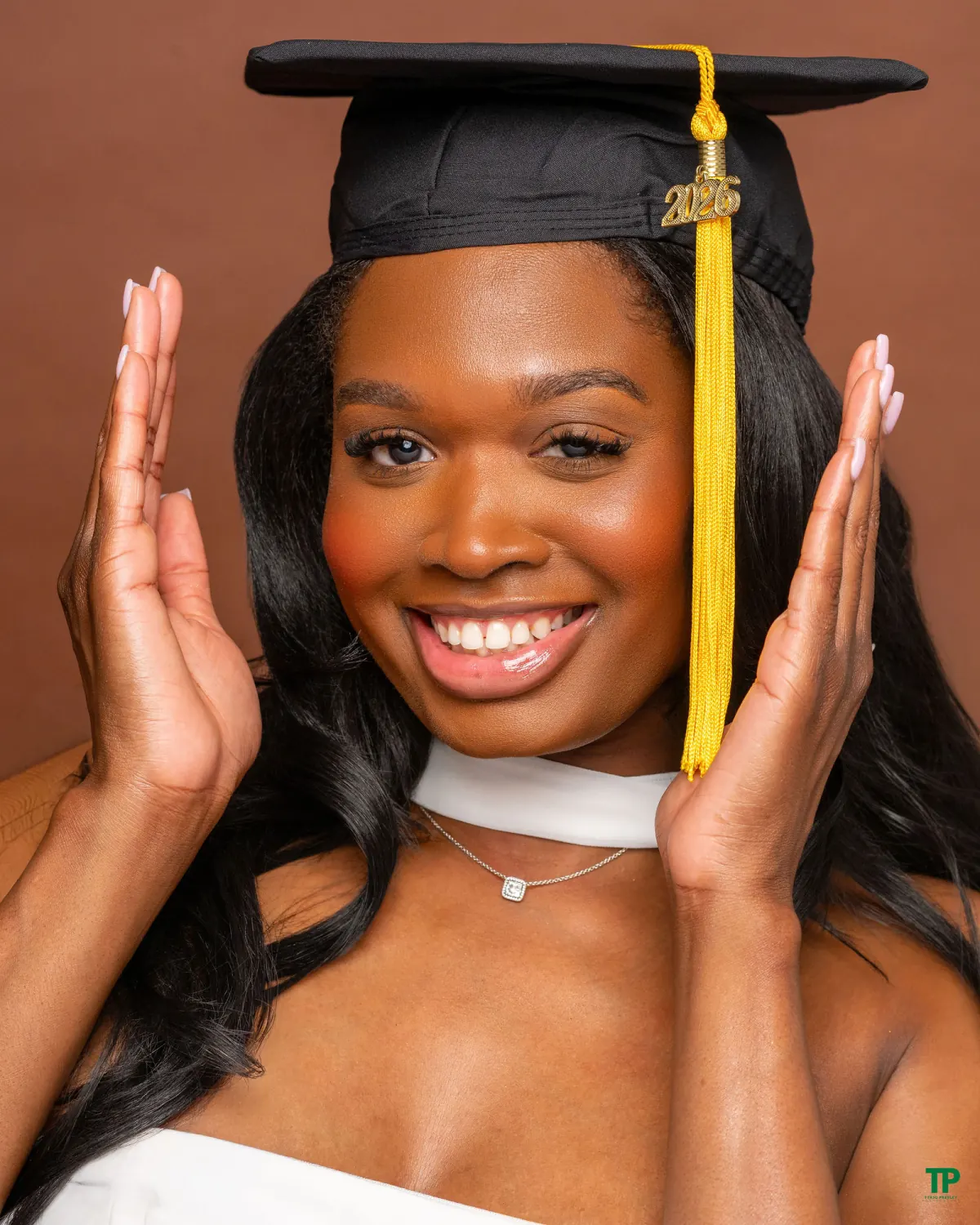 Smiling graduate in a white outfit holding her hands beside a black graduation cap against a warm brown background