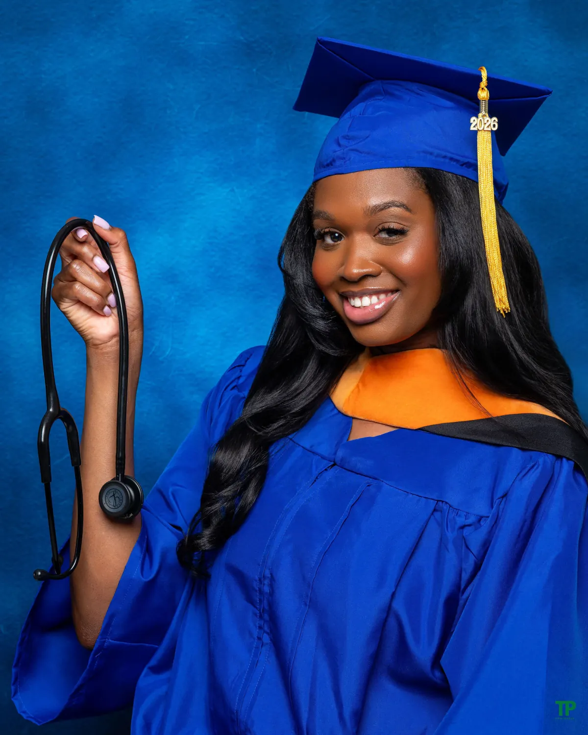 Smiling graduate in a blue cap and gown with an orange hood holding a stethoscope against a blue studio background