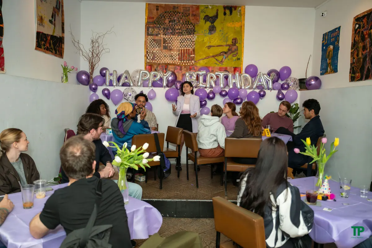 Guests gathered around tables listening to a speaker beneath purple birthday balloons