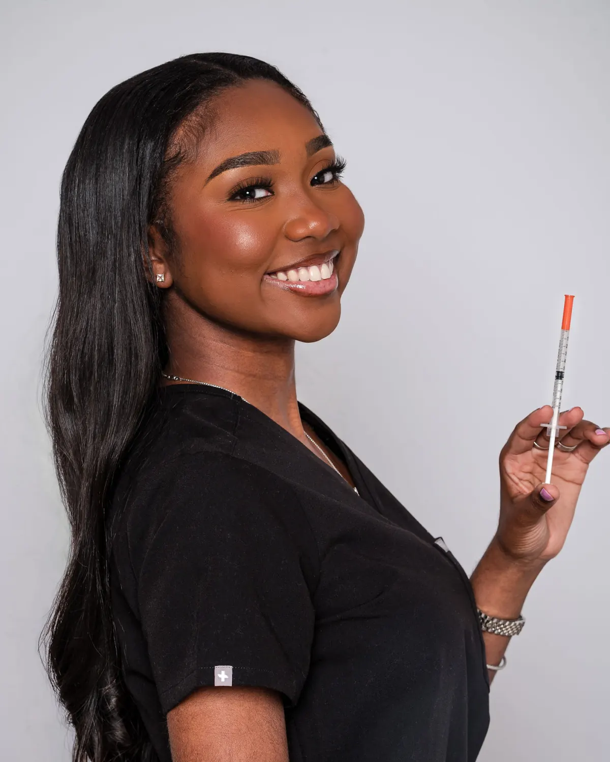 Smiling woman in black scrubs holding a capped syringe against a light studio background
