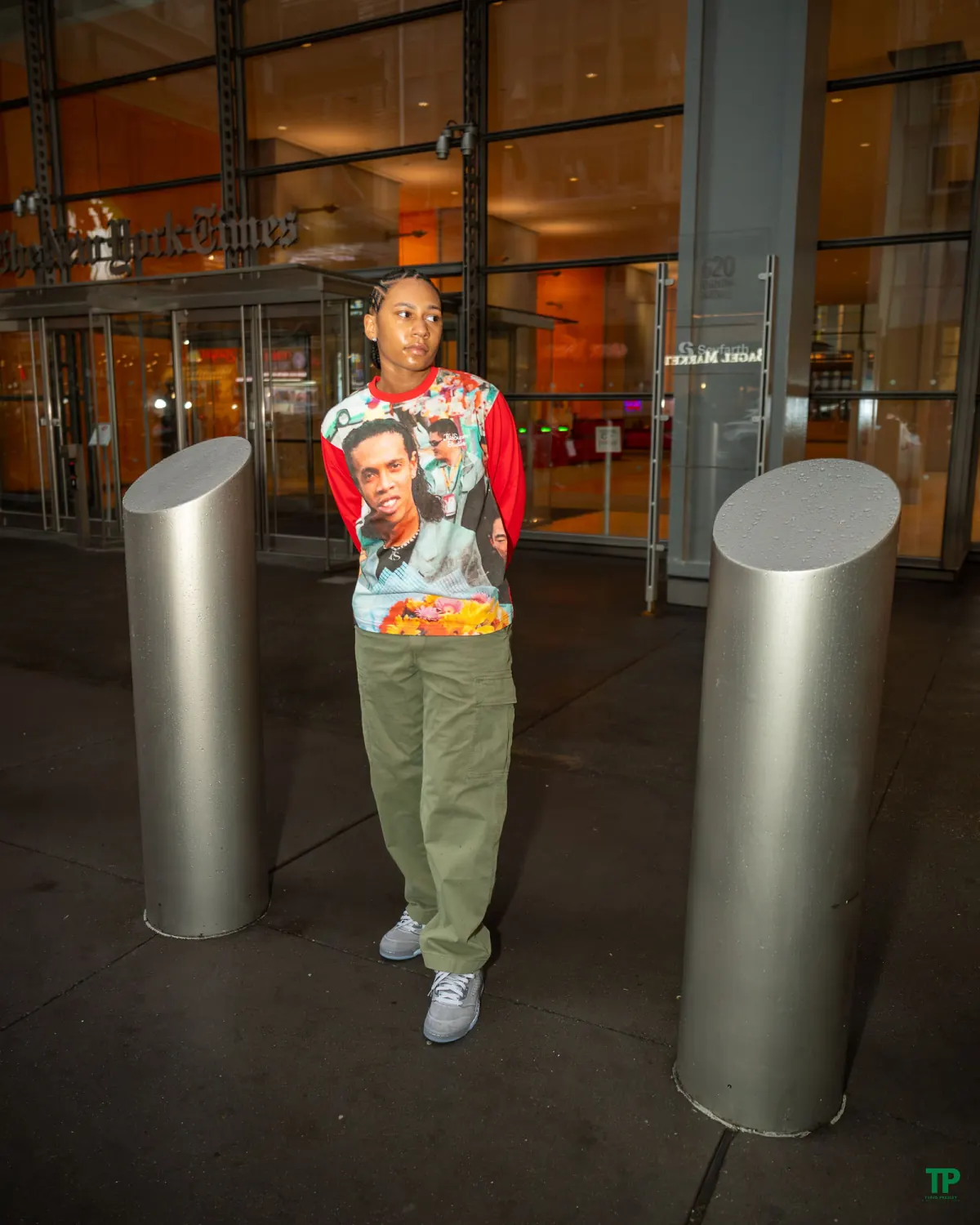 Person in a colorful red sweatshirt and green trousers standing between silver bollards outside a glass building at night