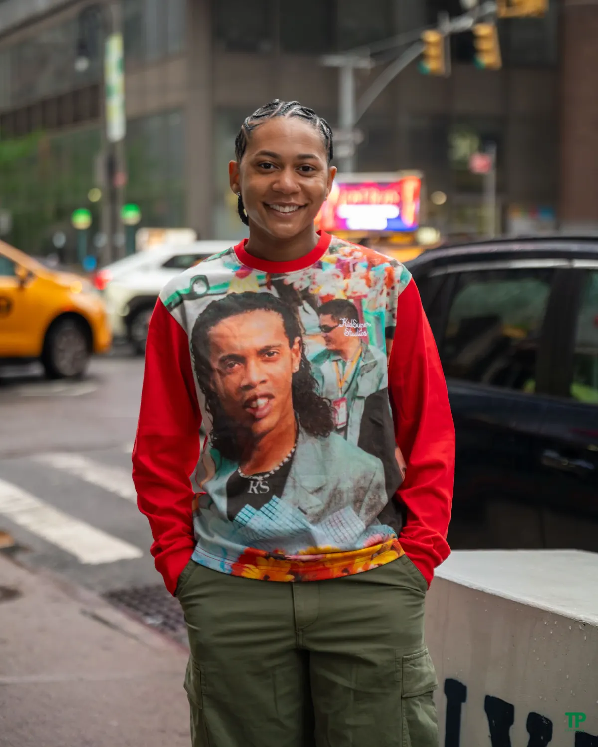 Smiling person with braids in a colorful red graphic sweatshirt on a city sidewalk